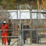 Um homem de pele média e uniforme de prisão laranja é transportado por guardas atrás de uma cerca de arame farpado.