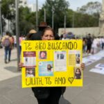 Uma mulher, Joanna Alvear, segura um cartaz amarelo brilhante, decorado com fotografias de sua filha desaparecida e fotos