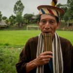 Tsonkiri, um ancião indígena, está em frente à sua aldeia amazônica com uma flauta de pã nos lábios.