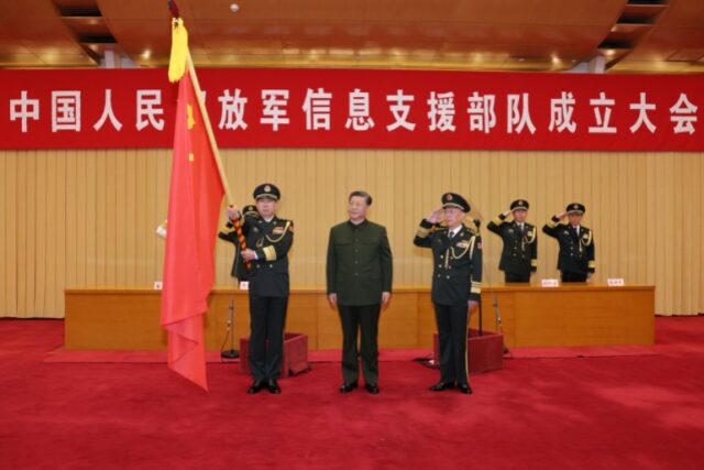 Xi Jinping apresenta uma bandeira à força de apoio à informação do ELP em abril.  Eles estão em um palco com tapete vermelho.  Xi está entre dois oficiais militares.  Há uma faixa atrás e dois soldados saudando ao fundo. 