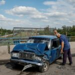 Um homem chora na porta de um carro azul onde seu filho foi morto em um ataque russo.  A frente do carro está destroçada com a placa pendurada.  O pára-brisa está quebrado.  Outro carro danificado está próximo.