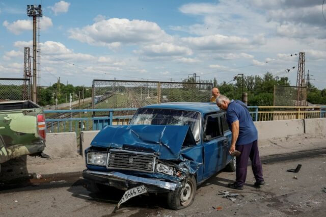 Um homem chora na porta de um carro azul onde seu filho foi morto em um ataque russo.  A frente do carro está destroçada com a placa pendurada.  O pára-brisa está quebrado.  Outro carro danificado está próximo.