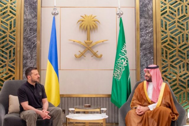 Volodymyr Zelenskyy em reunião com o príncipe herdeiro saudita Mohammed Bin Salman.  Eles estão sentados de cada lado de uma mesa de centro em frente às bandeiras de seus respectivos países.  Zelenskyy está com uma camisa pólo preta e calças cáqui.  Mohammed Bin Salman está com uma túnica tradicional. 