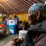 Uma mulher mais velha dentro de um gert tradicional da Mongólia.  Existe uma caixa de votação móvel configurada.  A mulher tem um lenço amarrado no cabelo e veste um casaco grosso.  Dois homens idosos estão na cabine de votação.  Há frascos e tigelas em uma mesa próxima.