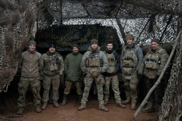 Soldados da brigada Azov posando para uma foto em janeiro de 2024. Eles estão diante de um canhão autopropelido perto da linha de frente.  Eles estão em uniformes de combate.