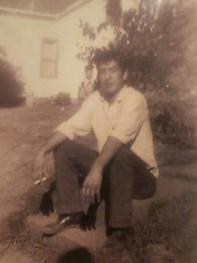 Uma foto em preto e branco de um jovem Leonard Peltier, sentado no chão do lado de fora, com uma camisa de colarinho branco e calça preta.