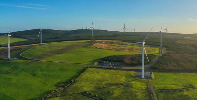 Planícies verdes repletas de grandes moinhos de vento.  Céu azul.