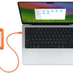 Vista aérea de um MacBook Pro aberto com um disco rígido externo laranja conectado (esquerda).