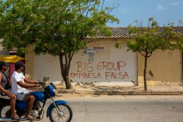 Dois homens em uma motocicleta passam por grafites que dizem em espanhol: 