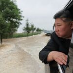 Kim Jong Un olhando para fora de um carro preto.  O veículo está sendo conduzido em águas marrons da enchente.  Kim parece sério.