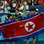 Jong Sik Ri da Coreia do Norte e Kum Yong Kim da Coreia do Norte seguram a bandeira da Coreia do Norte enquanto comemoram após a partida contra Chuqin Wang da China e Yingsha Sun da China