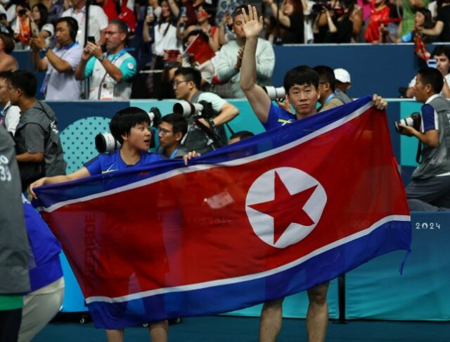 Jong Sik Ri da Coreia do Norte e Kum Yong Kim da Coreia do Norte seguram a bandeira da Coreia do Norte enquanto comemoram após a partida contra Chuqin Wang da China e Yingsha Sun da China