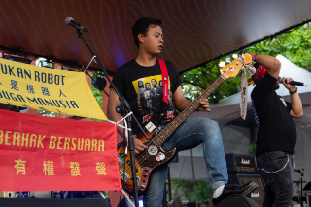 O guitarrista do Southern Riot tocando no palco.  Há faixas em indonésio que dizem “Não somos robôs” e “Também somos humanos”.  O guitarrista tem cabelos pretos cacheados e veste jeans e camiseta preta.  Ele está com o pé esquerdo apoiado em um dos alto-falantes e equilibrando o violão no joelho