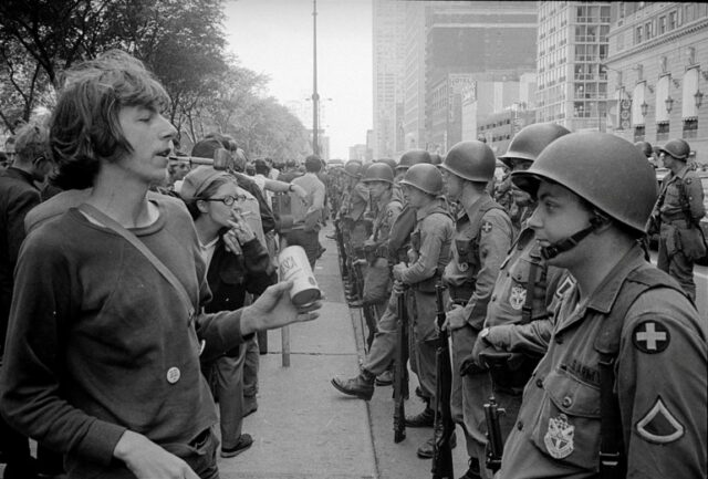 Uma foto de arquivo em preto e branco mostra policiais armados e com capacetes e manifestantes alinhados, frente a frente em uma ampla avenida de Chicago.