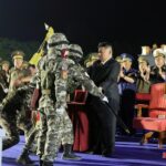 Kim Jong Un entregando uma bandeira a um soldado em uniforme de combate.  Eles estão no palco em uma cerimônia, Kim está vestida de preto.  Os soldados estão em uniforme de combate.  Existem outros funcionários por perto,