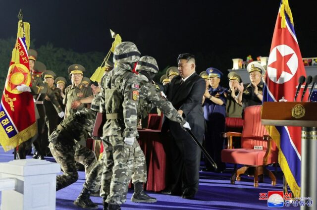 Kim Jong Un entregando uma bandeira a um soldado em uniforme de combate.  Eles estão no palco em uma cerimônia, Kim está vestida de preto.  Os soldados estão em uniforme de combate.  Existem outros funcionários por perto, 