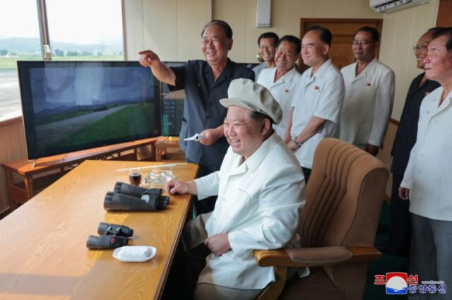 Kim Jong Un sentado em uma mesa. Há uma tela à sua frente mostrando a imagem de um tanque. Há dois pares de binóculos sobre a mesa. Kim está vestindo um jaleco branco e um boné bege claro na cabeça. Ele está rindo. Um grupo de homens mais velhos está com ele. Eles também estão sorrindo. Um está apontando.