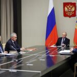 Vladimir Putin em reunião com autoridades governamentais.  Eles estão sentados a uma mesa.  Putin está à frente.  Há bandeiras atrás dele e um brasão na parede.