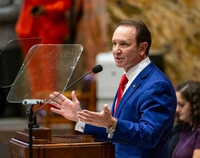 Jeff Landry fala atrás de um pódio de madeira que tem um escudo transparente na frente do microfone.  Ele veste um terno azul e gravata vermelha.