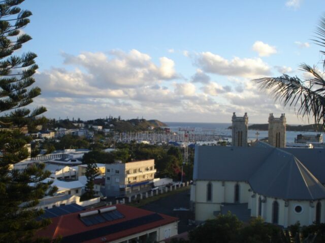 Uma vista de Noumea de uma colina acima da cidade.  O céu está azul com nuvens dispersas.  Parece tranquilo 