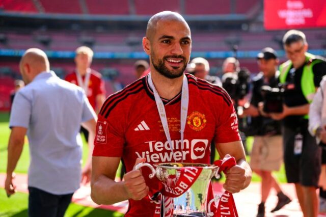 Amrabat segurando o troféu da FA Cup e vestindo um uniforme do Man United. 