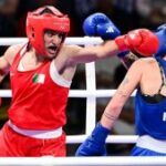 A boxeadora Imane Khelif veste vermelho, lançando um cruzamento contra seu oponente de azul no meio de um ringue de boxe, uma multidão sentada observando ao fundo.