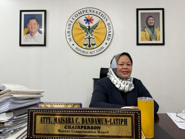 Maisara Dandamun-Latiph, ela está sentada em uma mesa com selos oficiais e um retrato do presidente na parede atrás dela. Ela está usando um vestido preto e um lenço branco na cabeça.