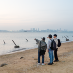 Três homens numa praia em Kinmen. Eles estão parados na areia apontando para Xiamen, que pode ser vista à distância. Existem picos anti-invasão na praia