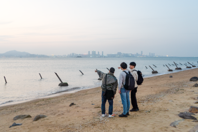 Três homens numa praia em Kinmen. Eles estão parados na areia apontando para Xiamen, que pode ser vista à distância. Existem picos anti-invasão na praia