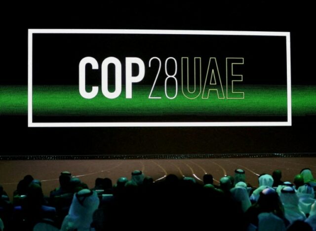 O logotipo 'Cop28 UAE' em branco é exibido na tela com um raio verde atravessando-o, público mal iluminado no meio da multidão em Abu Dhabi