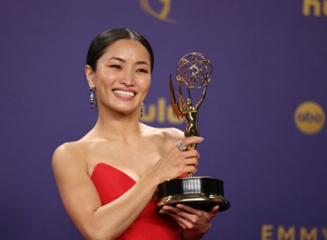 Anna Sawai segurando seu Emmy. Ela está usando um vestido de noite vermelho. Ela está sorrindo.