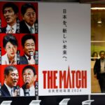 Um pôster gigante com retratos dos candidatos. Lê 'The Match' em inglês e japonês. Um homem está caminhando ao lado dele.