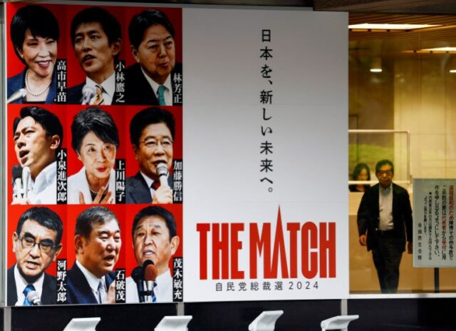 Um pôster gigante com retratos dos candidatos. Lê 'The Match' em inglês e japonês. Um homem está caminhando ao lado dele. 