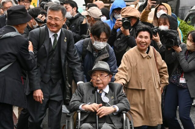 Iwao Hakamada comparece ao tribunal em Tóquio com sua família e apoiadores. Ele é uma cadeira de rodas. e usando óculos. Sua irmã está ao lado dele e sorri enquanto caminha.