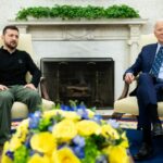 Volodymyr Zelenskyy e Joe Biden no Salão Oval. Eles estão sentados em frente a uma lareira. Há uma grande exposição de flores azuis e amarelas em uma mesa à frente deles.