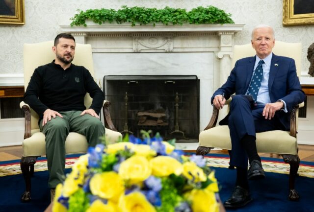 Volodymyr Zelenskyy e Joe Biden no Salão Oval. Eles estão sentados em frente a uma lareira. Há uma grande exposição de flores azuis e amarelas em uma mesa à frente deles.