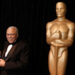 James Earl Jones segurando seu Oscar honorário. Ele está de pé contra um fundo preto. Há uma estatueta gigante do Oscar à direita da foto.