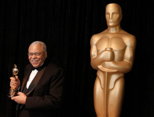 James Earl Jones segurando seu Oscar honorário. Ele está de pé contra um fundo preto. Há uma estatueta gigante do Oscar à direita da foto. 