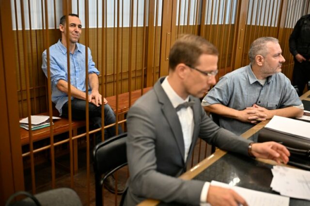 Laurent Vinatier sentado em uma doca em seu julgamento. Ele está vestindo uma camisa azul clara e calças escuras e com as mãos nos joelhos. Seus advogados estão sentados em uma mesa em frente à jaula