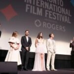 Um grupo de pessoas está em um palco; ao fundo, uma tela de cinema tem uma projeção onde se lê Festival Internacional de Cinema de Toronto. Há seis pessoas no palco, incluindo duas mulheres e quatro homens. Todos eles têm pele clara.