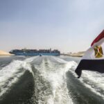 Uma bandeira nacional egípcia tremula em um barco enquanto mostra um navio porta-contêineres navegando na nova seção do Canal de Suez, na cidade portuária egípcia de Ismailia, 135 km a nordeste do Cairo, em 14 de outubro de 2019. - Desde que o Canal de Suez foi inaugurado em meio à pompa e cerimônia há 150 anos, tornou-se uma das vias navegáveis ​​mais importantes do mundo. Mas o seu aniversário só será comemorado discretamente no Egito.