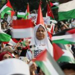 Uma mulher observa enquanto as pessoas levantam bandeiras palestinas durante um protesto contra Israel e em apoio aos palestinos do lado de fora da embaixada dos EUA em Jacarta, Indonésia, em 6 de outubro de 2024.